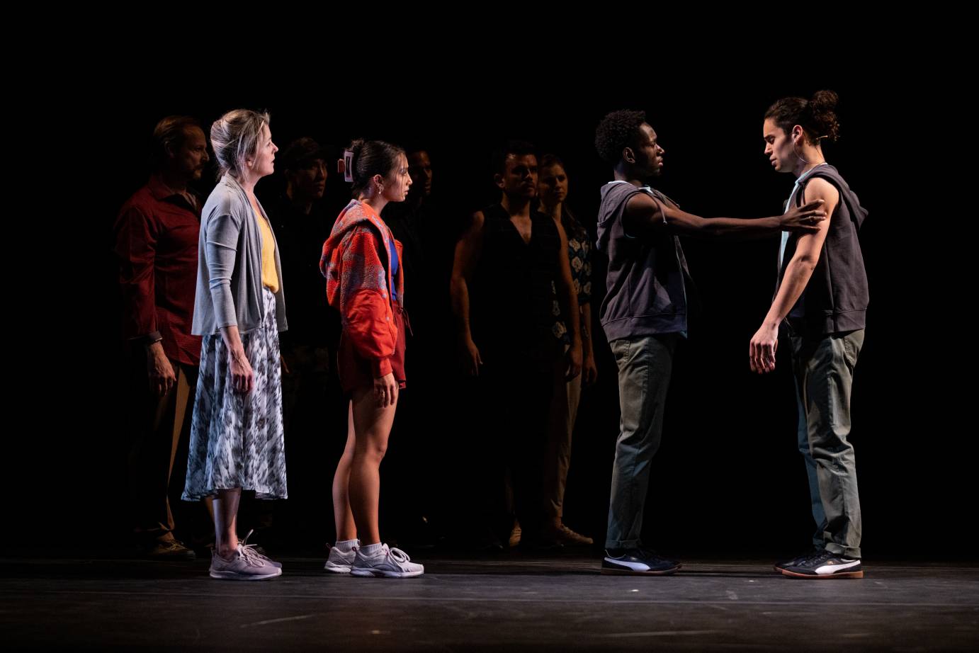 Nadia, in patterned skirt, yellow shirt, gray 'sweater', highlighted blonde hair, sneakers, stands next to Daria, athlete, dressed in white sneakers, augergine short shorts, blue top, red patterened hoodie. Both face stageleft and watch Felisberto meet his singer double, both dressed in sleeveless dark gray hoodie, sage green pants, sneakers. Felsiberto, the dancer, places his right hand on the upper left arm of singer, David Merino, who looks down